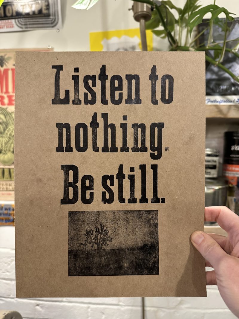 A hand holding up a print on chipboard with black ink. It says, "Listen to nothing. Be still." There is an electrotype plate of a joshua tree underneath the text.