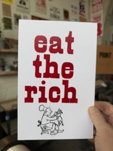A hand holding a sign that says, "eat the rich" with an illustration of a chef cooking a goose under it.