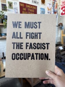 A hand holding a poster that says "WE MUST ALL FIGHT THE FASCIST OCCUPATION."