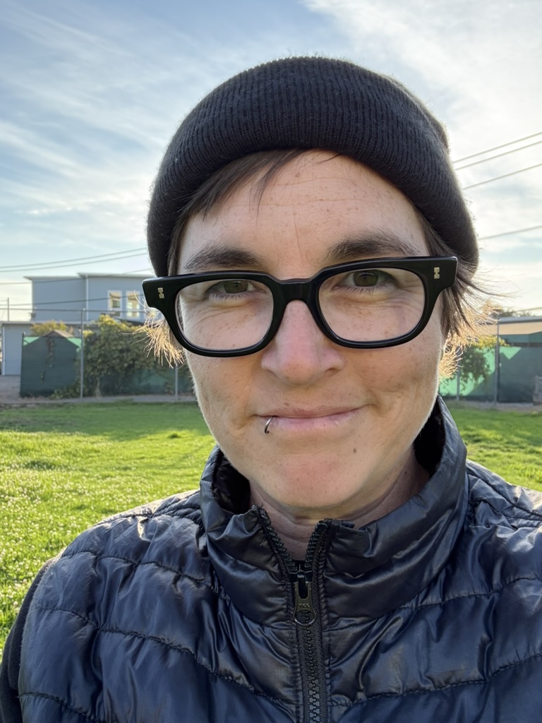 A middle aged person wearing a black toque, black glasses, and a black puffer vest. They are looking bemused.