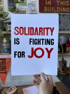 A hand holding a poster that says, "Solidarity it fighting for JOY"